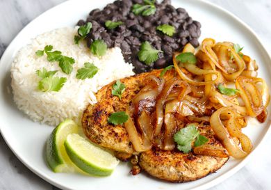 A plate of Cuban-style pollo a la plancha topped with caramelied onions, and served with white rice, black beans, lime wedges, and cilantro.