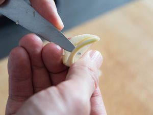 Hands holding a pairing knife and a halved garlic clove. The tip of the pairing knife is prying the germ from the clove.