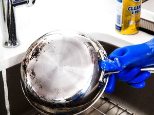 A blue-gloved hand holding a stainless steel skillet under a faucet