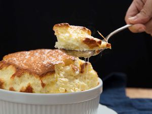 A large savory cheese souffle with someone holding a spoon of souffle above the casserole. 