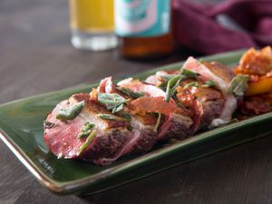 Sliced duck breast on a green rectangular platter, with a glass of sour beer in the background