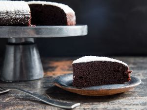 A slice of dairy-free chocolate cake served on a slate-colored plate. The remaining cake is nearby, perched on a stainless steel cake stand.