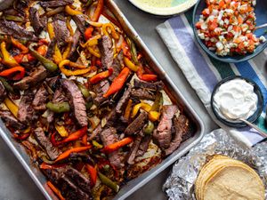 A sheet pan loaded with skirt steak strips, bell peppers, and onions, all after being cooked until charred, ready to be loaded into tortillas for fajitas