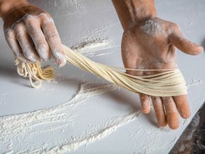 Hands holding thin hand-pulled lamian noodles over floured counter.