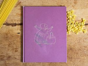 A copy of the book Strega Nona by Tomie dePaola, on a wood tabletop with dried pasta. .