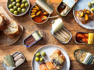 overhead shot of an array of tinned fish, or conservas, on a wooden table with bread and olives.