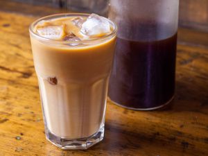 A glass container of cold brew coffee concentrate behind a full glass of iced coffee and milk.