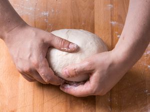 Two hands gently hold a smooth ball of dough on a wooden work surface.