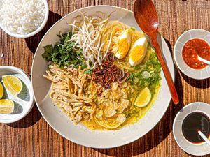 Bowl of Soto Ayam with wooden spoon, surrounded by limes, sambal, soy sauce and rice on a bamboo matt