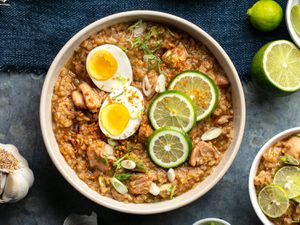 An overhead photo arroz caldo (Filipino chicken and rice soup), topped with a split boiled egg, lime wheels, and sliced scallions. A bulb of garlic and a halved lime is next to the bowl.