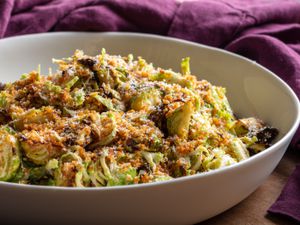 A bowl full of this salad, showing the varied textures of toasted breadcrumbs, thinly shaved raw Brussels sprouts, and crispy roasted ones, plus grated cheese, all lightly dressed in a creamy dressing.