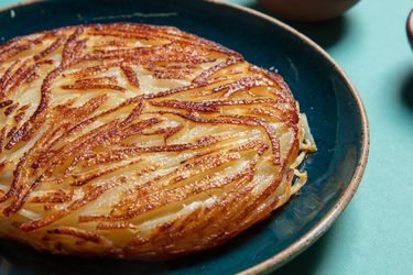 A golden brown potato pancake sits on a plate. It is round, thick, and is made up of clearly defined potato strands.