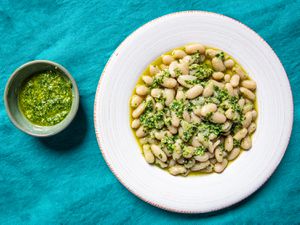 Overhead view of garlic scape pesto in a small bowl next to a wide rimmed, shallow bowl of cooked white beans and pesto