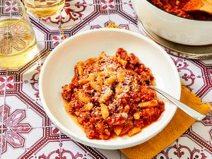 Overhead view of Malloreddus alla Campidanese in a white bowl on a titled background with pink floruishes surrounded by the pot and two wine glasses