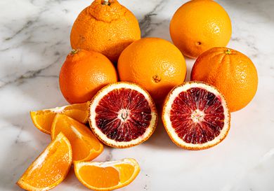 Whole and cut up oranges against a white marble countertop.