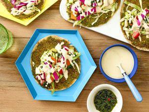 A smash falafel pita dressed with toppings next to more smash falafel pitas, a bowl of tahini sauce, and a bowl of zhug. 