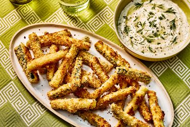 Baked Zucchini fries on a white dish, with a bowl of dip on the side. The table has green printed table cloth and two glasses of water. 