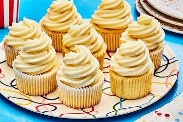 Horizontal image of 8 vanilla cupcakes on a colorful plate, and blue table. To the side is a stack of serving plates and colorful napkins