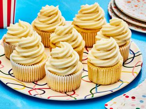 Horizontal image of 8 vanilla cupcakes on a colorful plate, and blue table. To the side is a stack of serving plates and colorful napkins