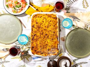 Pastel De Choclo in white dish, on a tabletop with plates, glasses of water and wine, and a salad on the side 