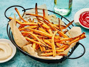 Basket of sweet potato fries on a blue surface, with small dishes of ketchup and aoli sause, and a bottle of water