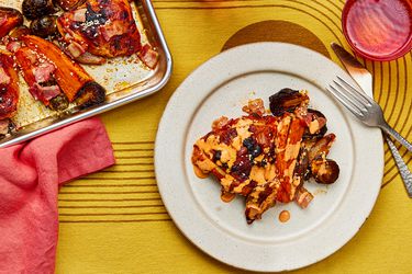 Serving of sheet-pan chicken and vegetables on a plate with a fork and knife and a red cup. To the side of the dish is the tray of chicken and vegetables with a red napkin