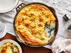 Baked Pot pie with one serving taken out of skillet and on a plate. Skillet is on a white tablecloth and has serving plates around it. 