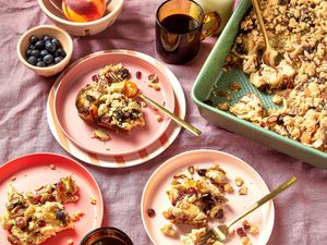 Challah Breakfast bake servings on 3 different plates, with a forks, coffee, milk and tray of challah bake, blueberries and peaches in the background on a pink textile