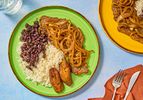 Green Plate of Pallomilla with rice beans and plantains. Red napkin with untensils, and large plate of pallomilla to the side 