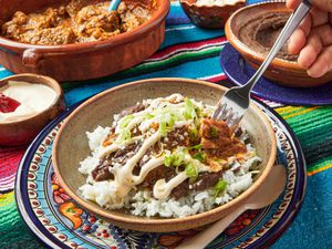 Dish of morisqueta on white rice, with a fork poking into dish. Dishes of white rice, water, and platter of moriqueta on the side. Table cloth is colorful Mexican striped textile