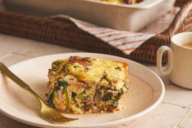 Serving of baked casserole square from the side on a plate with a gold fork. Coffee and basket of bigger casserole are in the background. 