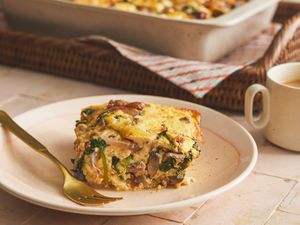 Serving of baked casserole square from the side on a plate with a gold fork. Coffee and basket of bigger casserole are in the background. 