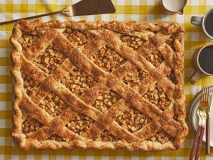 Whole Apple Slab Pie being placed on a plate. Tray is on a yellow gingham print tablecloth, with two cups of coffee, plates and gold forks 