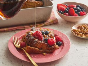 Gif of syrup pouring ontop of french toast casserole with berries, on a pink plate, with topping and casserole in the background