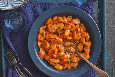 Glazed carrots in a blue bowl on blue rustic surface. A spoon is in the bowl, with a few carrot slices in it, and there is cups of sparkling water, and plates on the side.