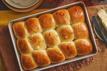 Tray of Parkerhouse Rolls, on a brown textile, with a side dish of butter and small serving dishes to the side. 