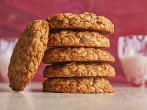 oatmeal raisin cookies stacked up on top of each other, and one cookie leaning on the stack. 2 glasses of milk in back and a red swirl background wall.