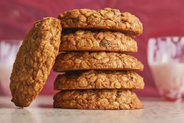 oatmeal raisin cookies stacked up on top of each other, and one cookie leaning on the stack. 2 glasses of milk in back and a red swirl background wall.