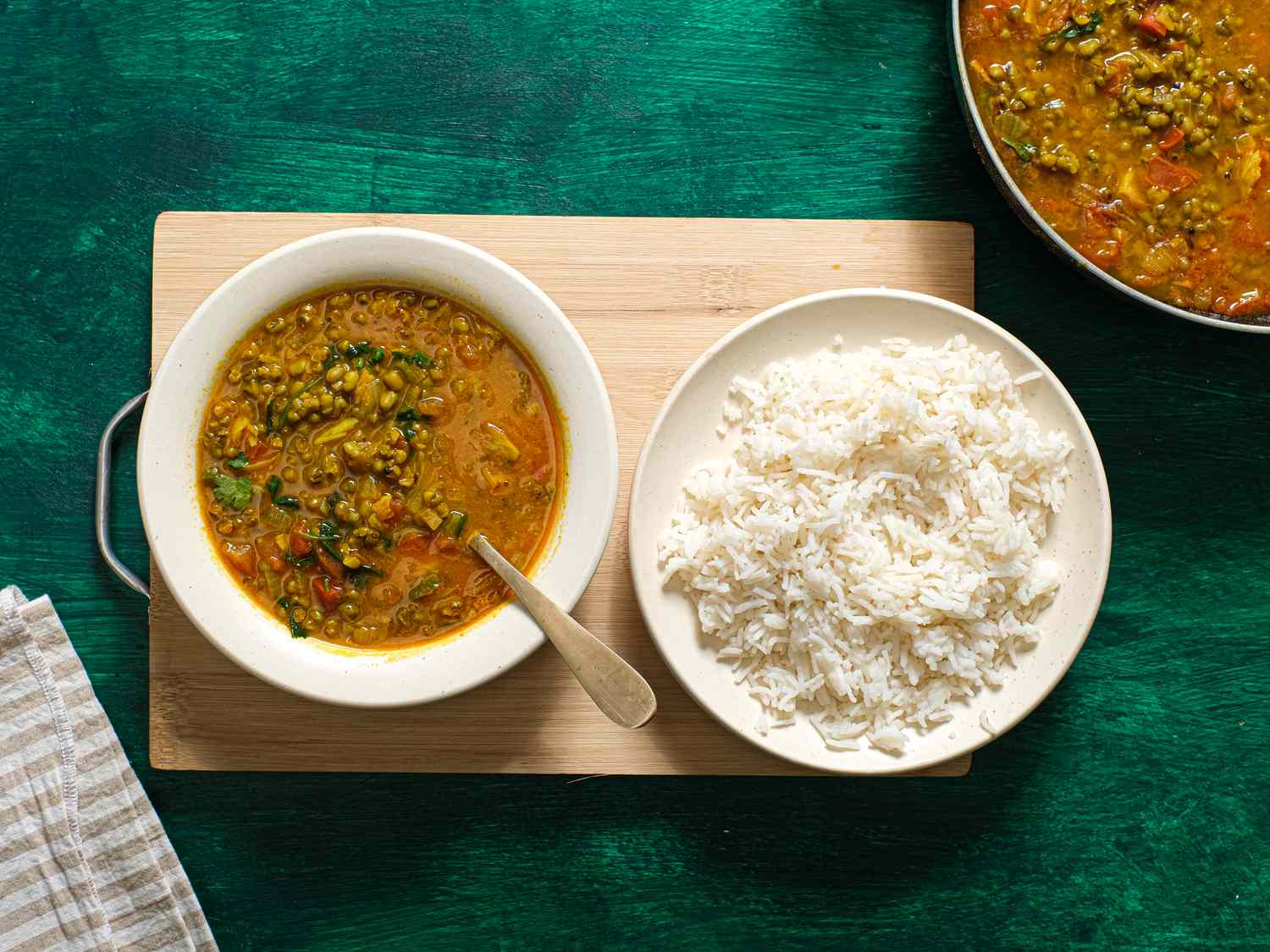 Bowl of Moong dal in a bowl, with a bowl of white rice next to it. Bowls are on a wooden board on top of green textured surface, and larger bowl of Dal is in the side of the frame. 