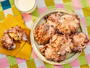 Platter of apple fritters on a round dish, with one fritter on yellow napkins, ripped open. Glass of milk, and colorful checkered tablecloth