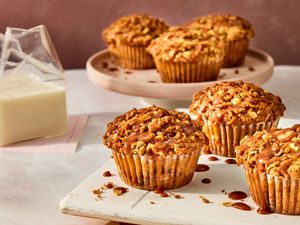 apple pie muffins on a white tray, with 3 muffins on a circular tray in the background. A clear glass of milk to the side on pink napkins. 