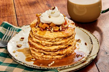 Stack of apple pie pancakes on white rustic plates, a green checked napkin, and warm drink