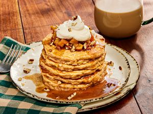 Stack of apple pie pancakes on white rustic plates, a green checked napkin, and warm drink