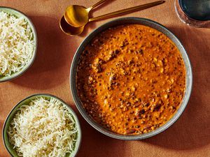 Dal Makhani in a bowl with 2 gold spoons, and 2 bowls of rice. Burnt orange colored surface and blue glass of water. 