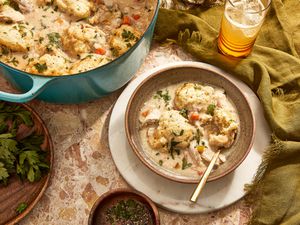 bowl of chicken dumpling soup on a rustic stoneware bowl, with a tarazzo tabletop, dark green textiles and the pot of soup to the side