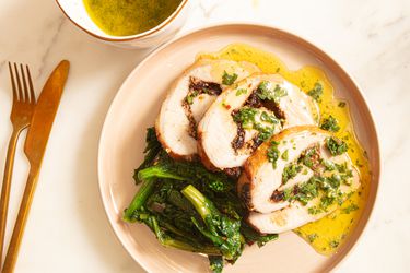 Stuffed pork loin slices with sauce sauce and greens on a warm colored plate. Gold fork and knife to the side and small bowl of sauce in top corner 