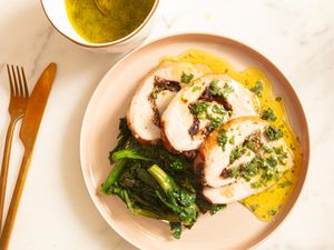 Stuffed pork loin slices with sauce sauce and greens on a warm colored plate. Gold fork and knife to the side and small bowl of sauce in top corner 