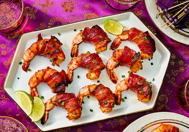 Bacon-wrapped shrimp lined up on a platter, with pink asian inspired fabric, and smaller plates and drinks around the sides