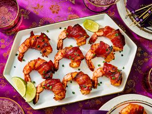 Bacon-wrapped shrimp lined up on a platter, with pink asian inspired fabric, and smaller plates and drinks around the sides