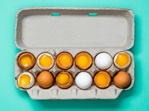 An open carton of a dozen eggs. Some of the eggs are cracked open so the yolks are exposed.